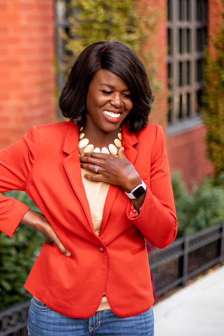 Atima Omara in bright orange jacket with hand to necklace.