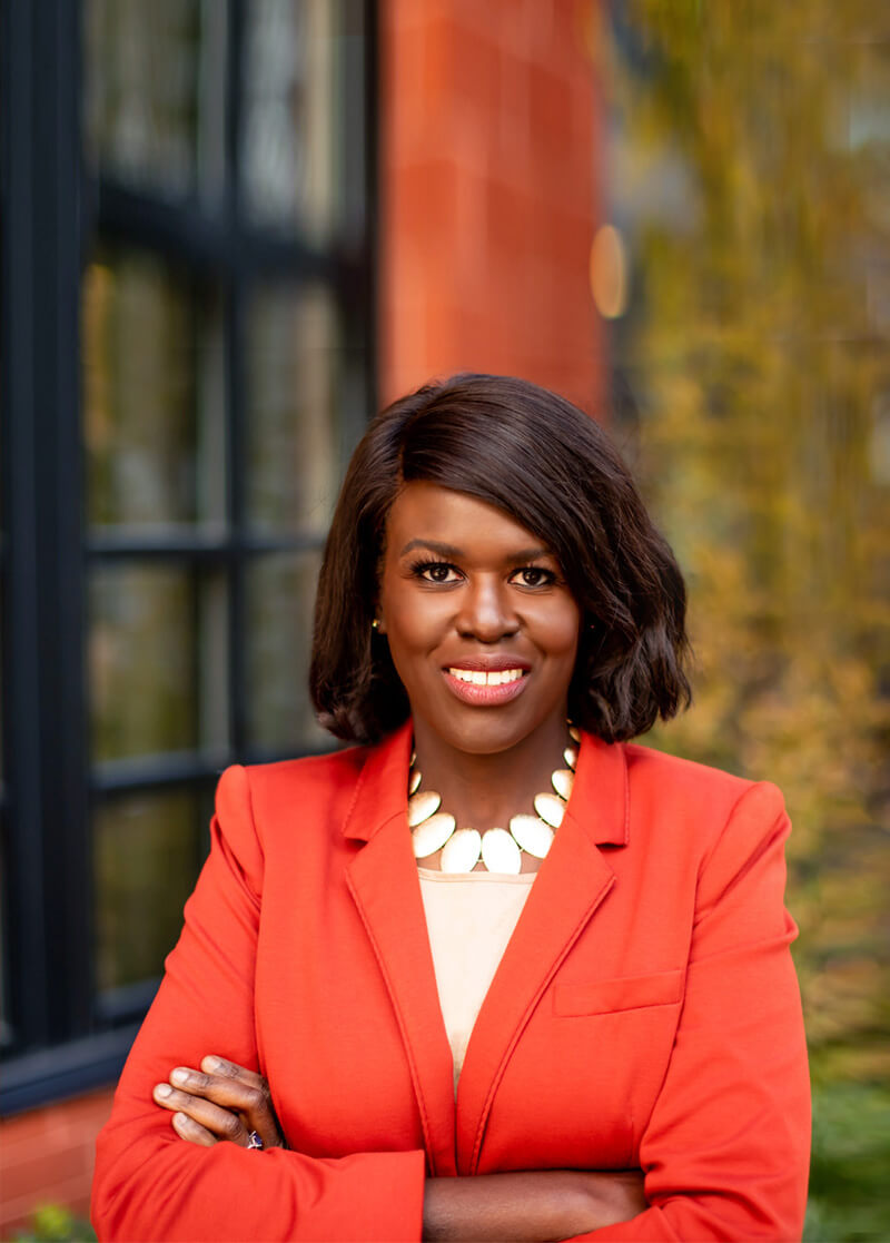 Atima Omara in bright orange jacket with arms crossed smiling, background blurred and cropped.